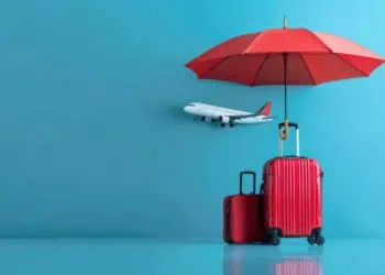Two red suitcases and a miniature-sized airplane appear in front of a blue background under a red umbrella signifying travel insurance