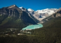 Aerial view of a turquoise lake surrounded by dense forest and majestic snow-capped mountains under a clear blue sky.