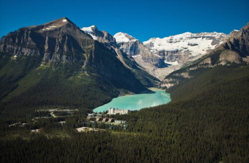 Aerial view of a turquoise lake surrounded by dense forest and majestic snow-capped mountains under a clear blue sky.