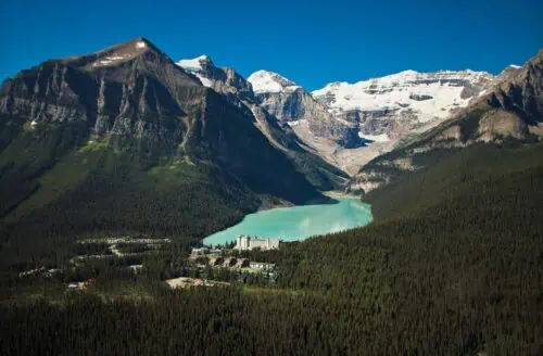 Aerial view of a turquoise lake surrounded by dense forest and majestic snow-capped mountains under a clear blue sky.