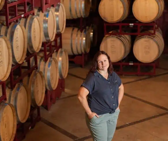 A woman stands in front of a large rack filled with barrels, showcasing a rustic storage area.